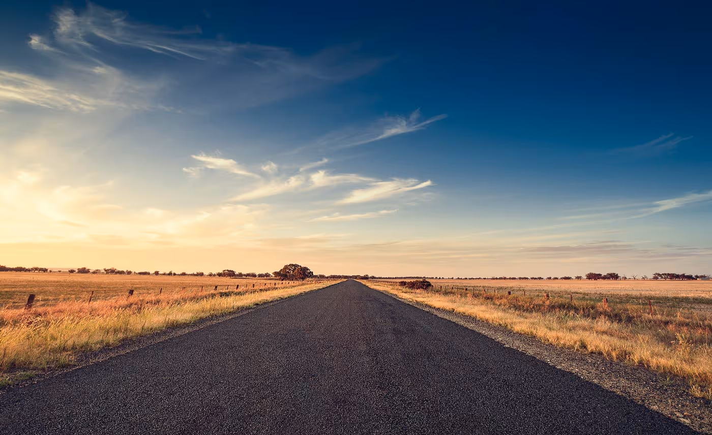 A straight, empty road stretches through flat, open fields under a blue sky.